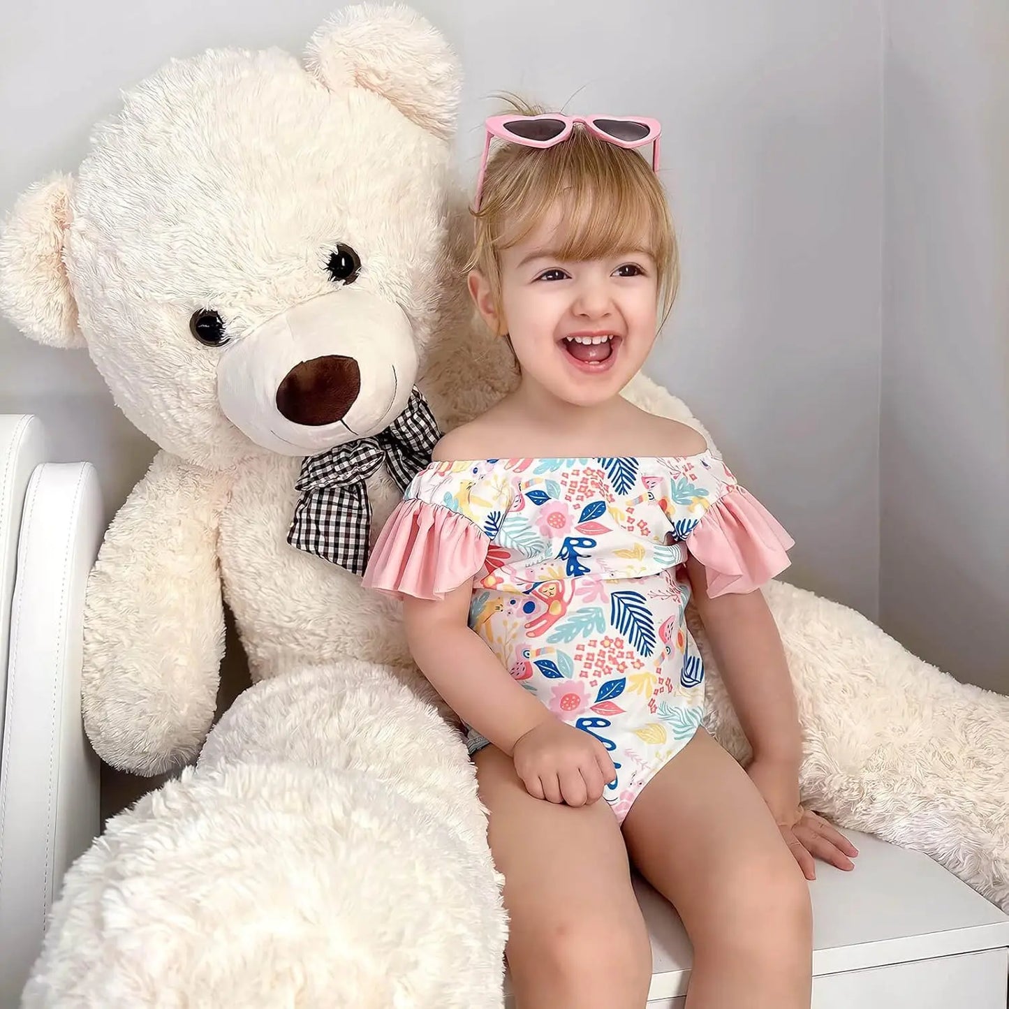 Child in a floral swimsuit with pink sunglasses sitting next to a large white teddy bear.