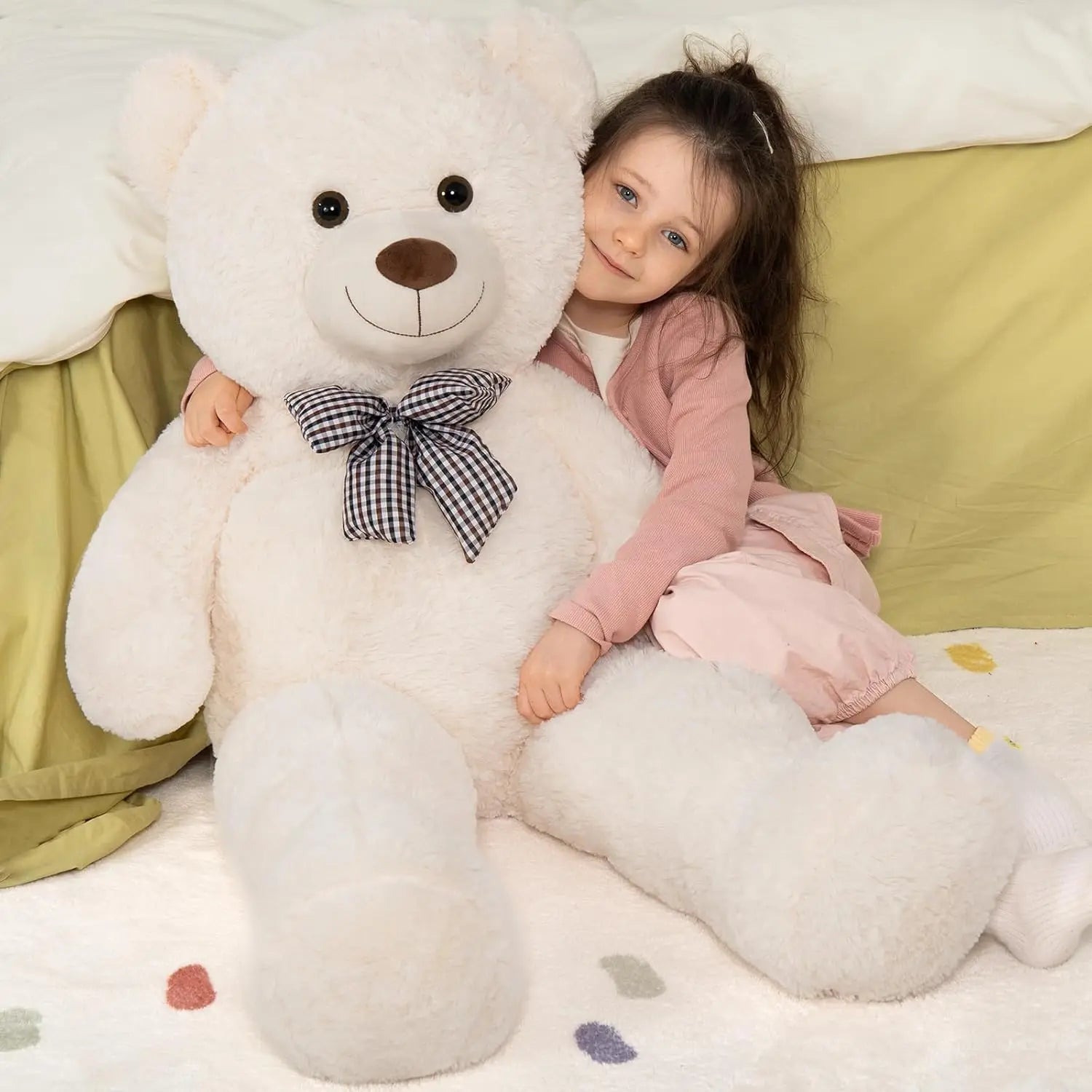 Child sitting on a large white teddy bear with a checkered bow, on a light green couch.