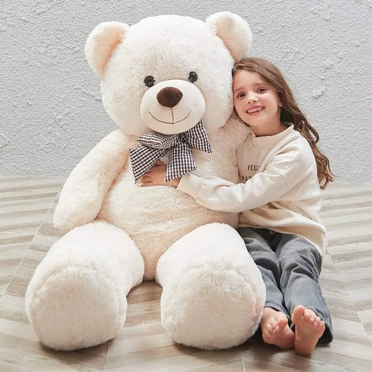 Child hugging a large white teddy bear with a checkered bow tie on a light wooden floor.