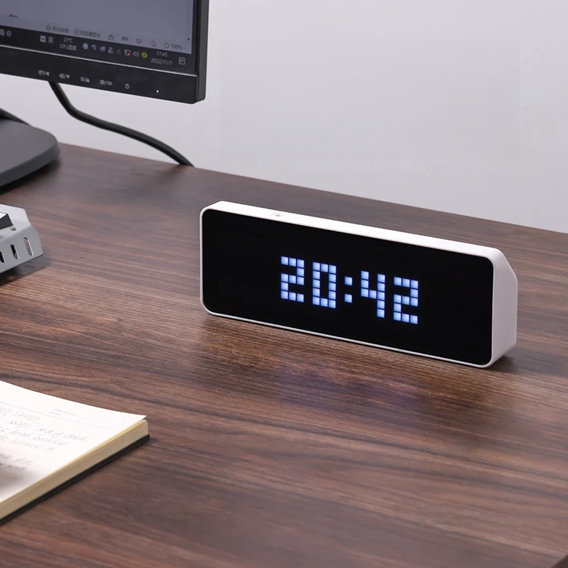 Digital clock displaying time on a wooden desk with a computer monitor in the background