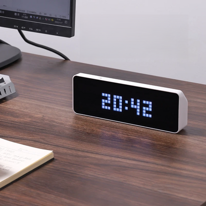 Digital clock displaying time on a wooden desk with a computer monitor in the background